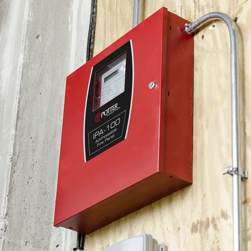 A red Potter IPA-100 addressable fire alarm panel mounted beside metal conduit
